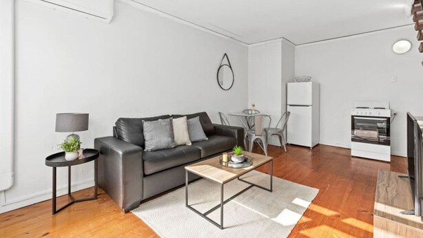 Un salon moderne avec un canapé en cuir noir, une petite table ronde avec une lampe, une table basse et un coin cuisine équipé d’une cuisinière et d’un réfrigérateur.