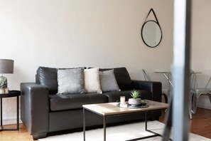 A modern living room with a black leather sofa, a small wooden coffee table, and a side table with a lamp and a plant. - Domi Rentals - The Murra Apartments (Murrumbeena)