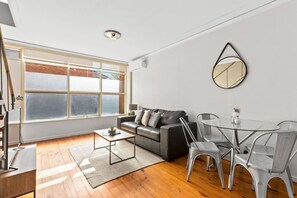 A modern living room with a sofa, a glass coffee table, and a dining table with chairs. - Domi Rentals - The Murra Apartments (Murrumbeena)