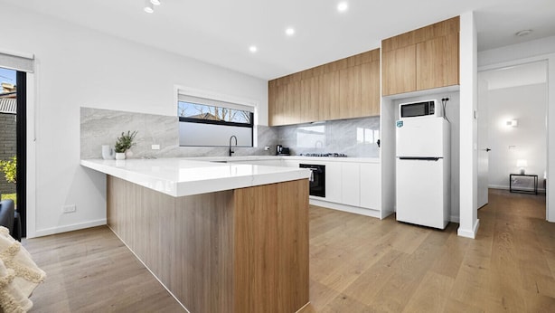 A modern kitchen with a central island, white cabinetry, and a view of the outdoors. - Domi rentals - The Highland House (Oakleigh East)