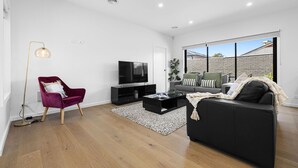 A modern living room with a black sofa, a red chair, a TV, and a large window. - Domi rentals - The Highland House (Oakleigh East)