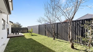 A residential backyard with a wooden deck, a small patio table, and a black fence. - Domi rentals - The Highland House (Oakleigh East)