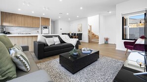 A modern living room with a black sofa, a coffee table, and a kitchen area with wooden cabinets. - Domi rentals - The Highland House (Oakleigh East)
