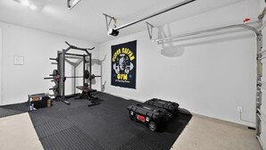 A home gym with a squat rack, bench, and weight plates; a banner on the wall; and a ceiling-mounted pull-up bar. - Modern 4BR, Sleeps 10, King Suite, Home Office, Garage Gym & Game Room (Manor)