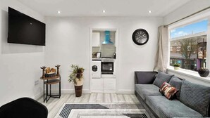 A modern living room with a sofa, a flat-screen TV, a kitchen area, and a clock on the wall. - Belvedere 4-Bed Apt, Sleeps 9 with Parking (Belvedere)