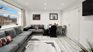 A modern living room with a sectional sofa, a dining table with chairs, a flat-screen TV mounted on the wall, and a large window providing a view of the outside. - Belvedere 4-Bed Apt, Sleeps 9 with Parking (Belvedere)
