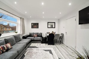 Ein modernes Wohnzimmer mit einer Eckcouch, einem Esstisch mit Stühlen, einem flachbildfernseher an der Wand und einem großen Fenster mit Blick ins Freie.