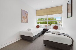 A bedroom with two beds, a window with a view, and a painting on the wall. - Domi Rentals - The Meadow Apartment (Saint Kilda East)