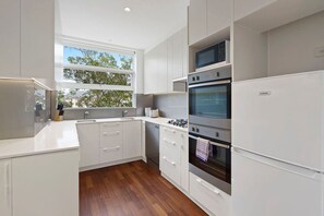 A modern kitchen with white cabinetry, a built-in oven, a microwave, and a window providing natural light. - Domi Rentals - The Meadow Apartment (Saint Kilda East)