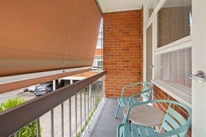 A balcony with a metal railing, a brick wall, and a window with curtains. - Domi Rentals - The Meadow Apartment (Saint Kilda East)