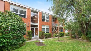 A multi-story residential building with a brick facade, a small front garden, and a bench.