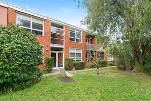 A multi-story residential building with a brick facade, a small front garden, and a bench. - Domi Rentals - The Meadow Apartment (Saint Kilda East)