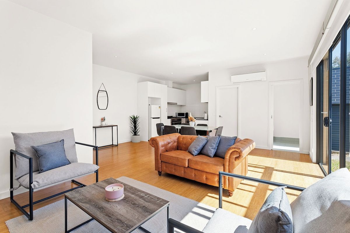 A modern living room with a sofa, armchair, and coffee table. A kitchen is visible in the background with white cabinets and a dining area.