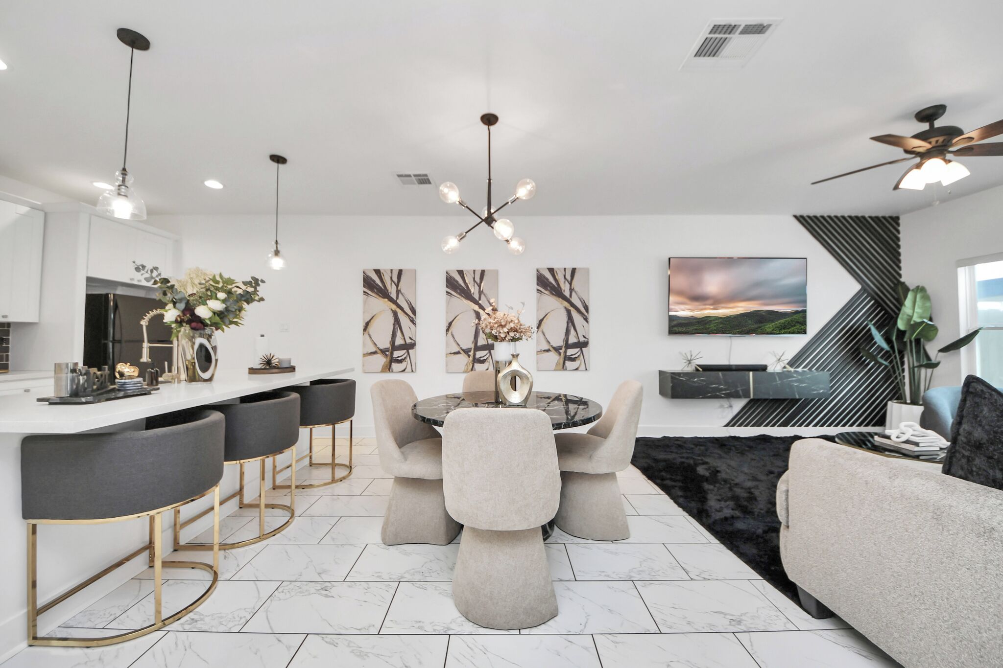 A modern kitchen with a dining area, featuring a central round table with chairs, a bar area with stools, and a large flat-screen TV mounted on the wall.