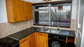 A kitchen with wooden cabinets, a sink, a stove, a washing machine, and a window with blinds. - Manchester City Short Let (Manchester)