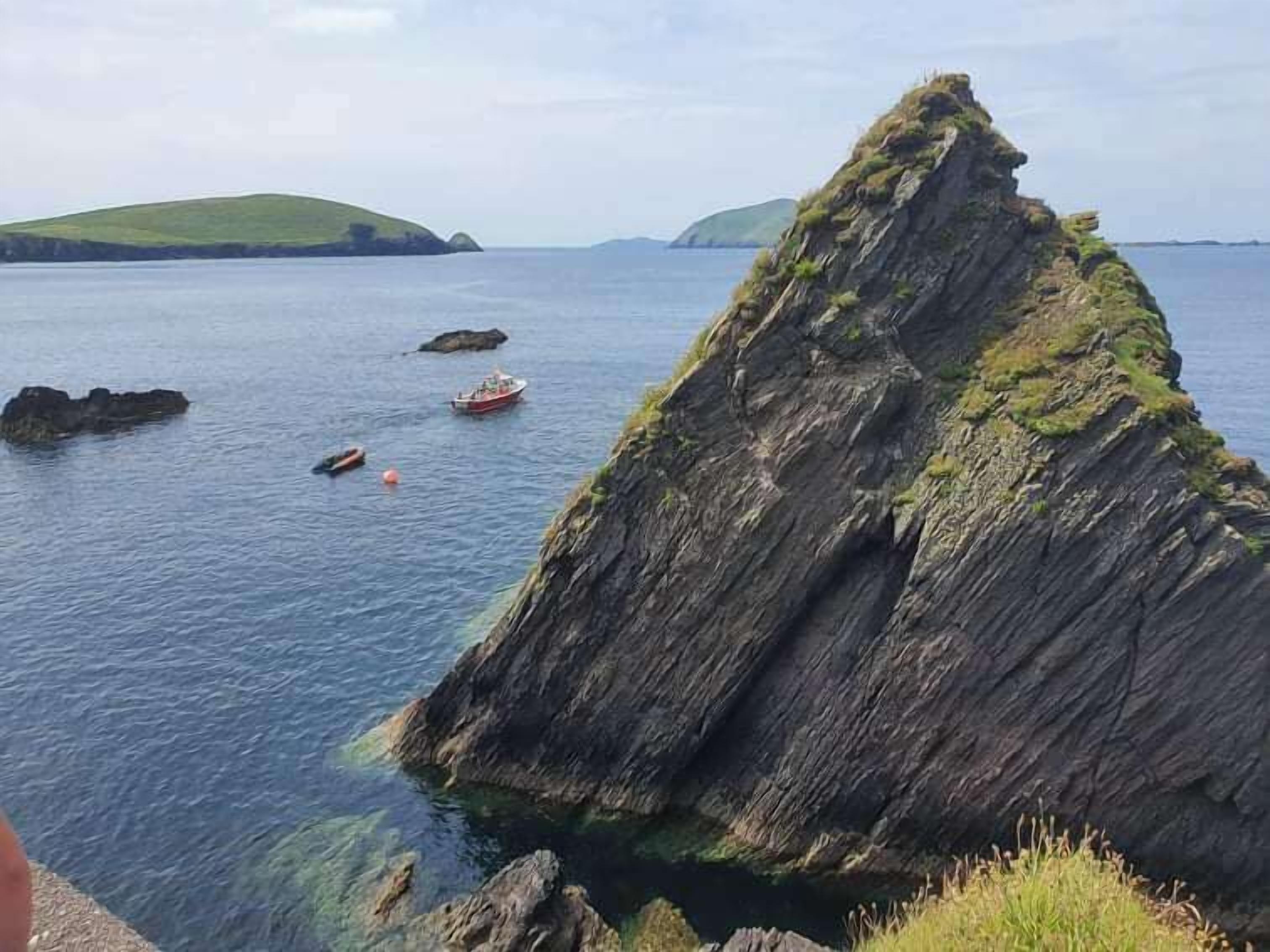 A coastal landscape with a prominent rock formation, calm sea, and a distant green hill.
