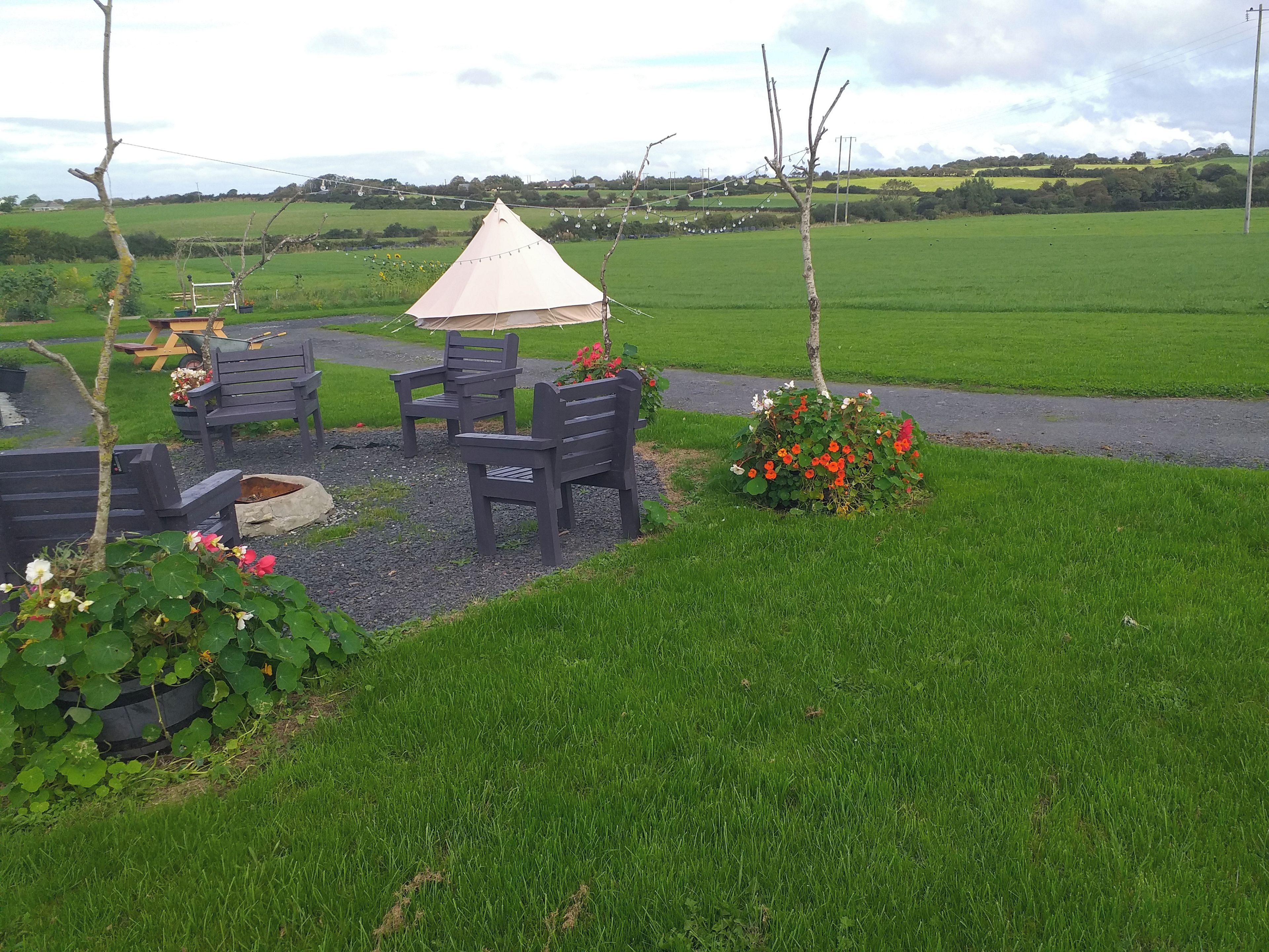An outdoor seating area with a table and chairs, a small garden with flowers, and a view of a green field.