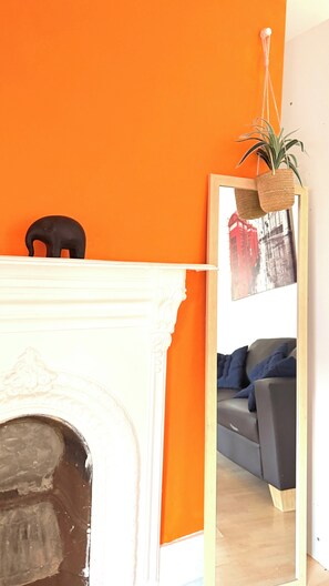Ein Zimmer mit weißem Kamin, einer orangefarbenen Wand, einem grauen Sofa und einem Spiegel, der einen Stadtblick reflektiert.