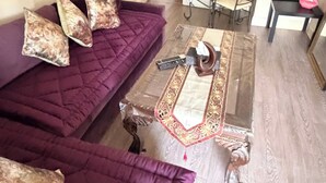 A living room with a purple sofa set, a glass coffee table, and a dining area with a metal chair and table. - Apartment Sleeps 6 with in Kilburn Lane (London)