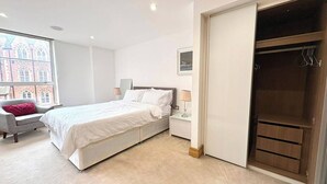 Ein Schlafzimmer mit einem Bett, einem Nachttisch, einem Sessel, einem Fenster mit Blick auf Gebäude und einem Kleiderschrank.