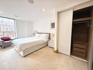 Ein Schlafzimmer mit einem Bett, einem Nachttisch, einem Sessel, einem Fenster mit Blick auf Gebäude und einem Kleiderschrank.