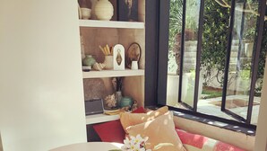 A room with a white round table, a vase with flowers, a white pitcher, and a bookshelf with various decorative items. - Bronte garden apartment (Waverley)