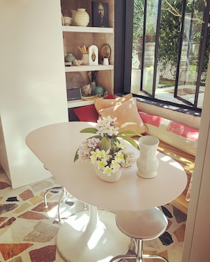 A room with a white round table, a vase with flowers, a white pitcher, and a bookshelf with various decorative items. - Bronte garden apartment (Waverley)