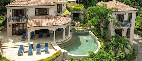 Et stort hus med swimmingpool, overdækket terrasse og flere liggestole.