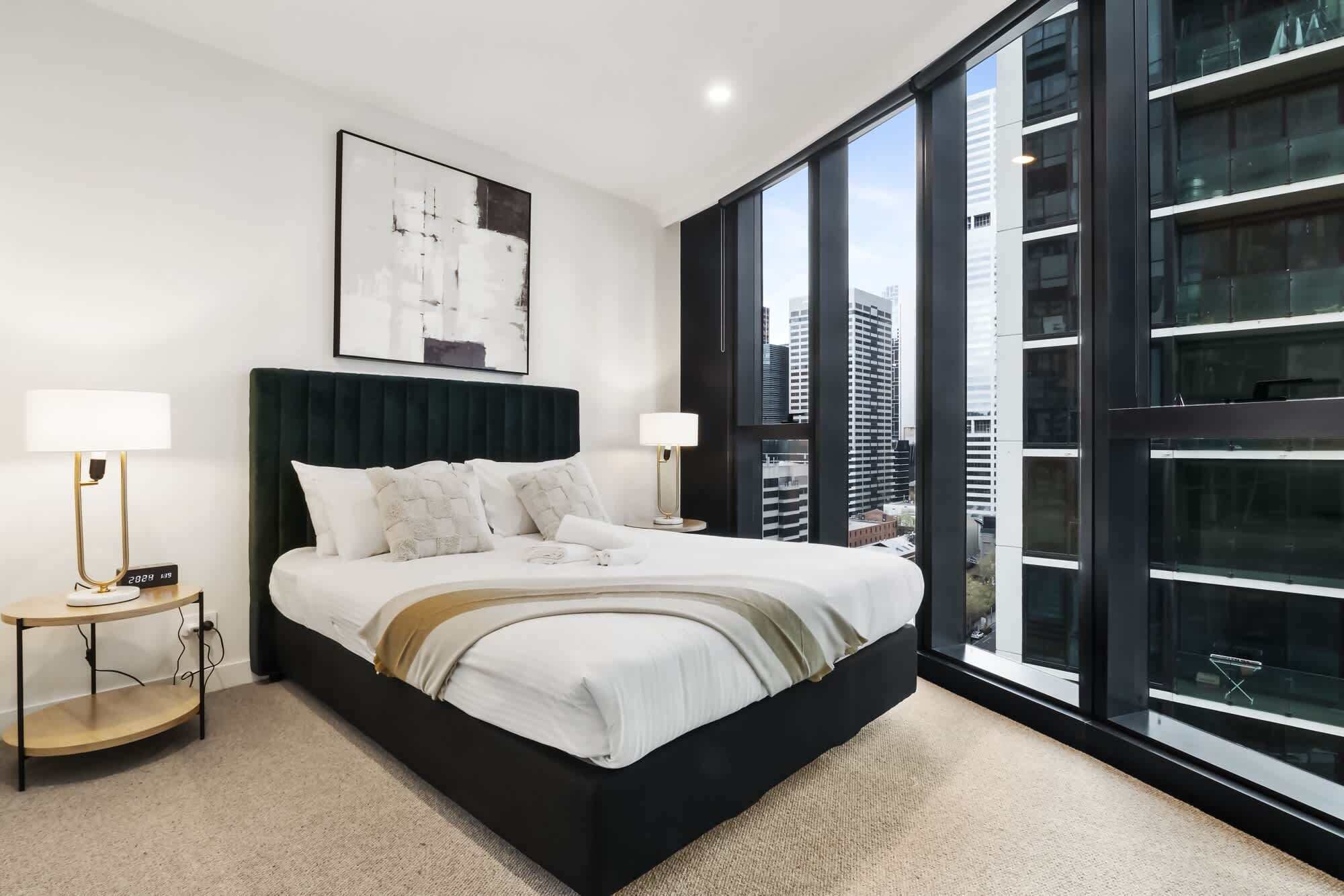 A modern bedroom with a large bed, two bedside tables, a nightstand, a wall-mounted clock, and a large window offering a city view.
