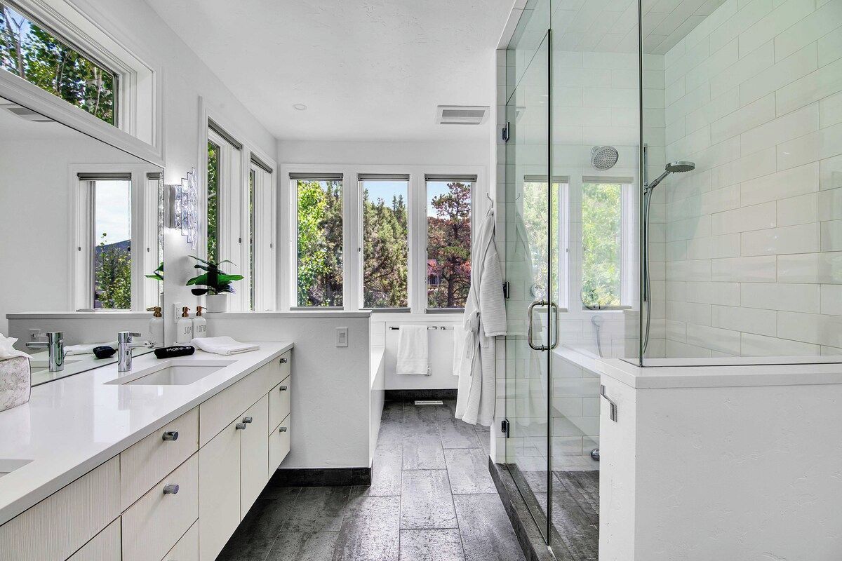 Un baño moderno con cabina de ducha de vidrio, un amplio ventanal con vista a árboles y un tocador blanco con espejo.