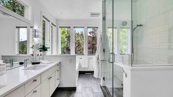 Un baño moderno con cabina de ducha de vidrio, un amplio ventanal con vista a árboles y un tocador blanco con espejo.