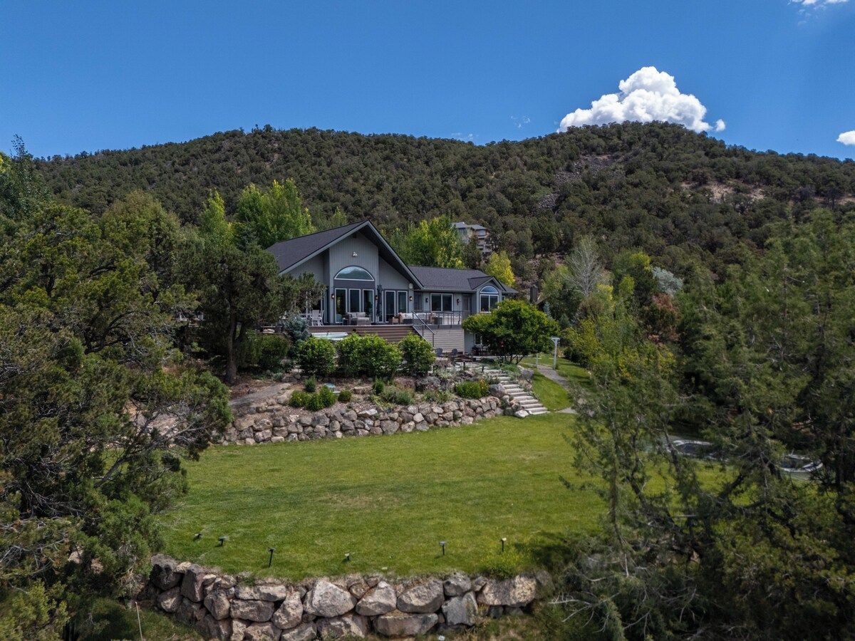 Una casa de dos pisos con un muro de contención de piedra, un porche y vista a un cerro boscoso.