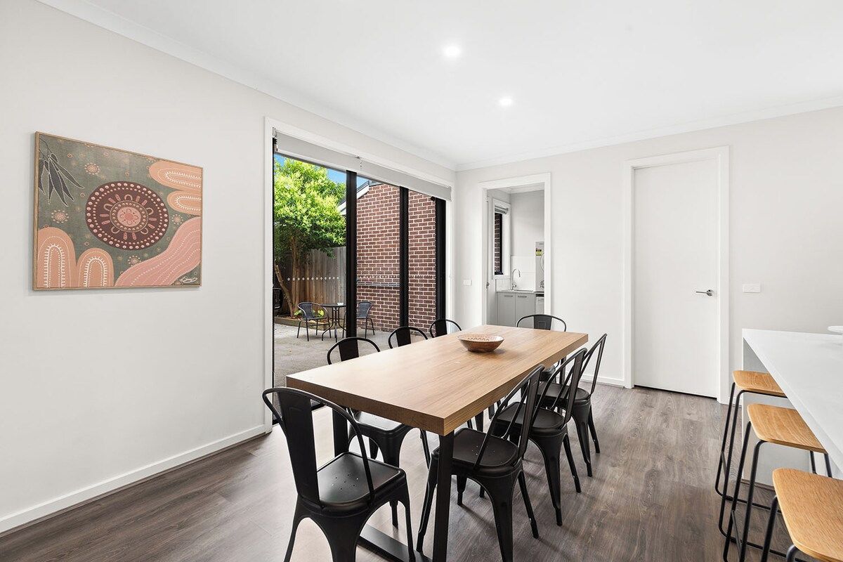 A modern dining area with a wooden table and black chairs, a white wall with a large abstract painting, and a view of a brick building outside.