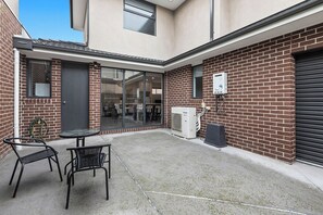 A courtyard with a brick wall, a glass door entrance, a table with chairs, and an air conditioning unit.
