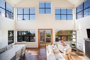 Un interior espacioso con techo alto, ventanas grandes, puerta corrediza de vidrio, un sofá blanco, una mesa de comedor y un televisor.