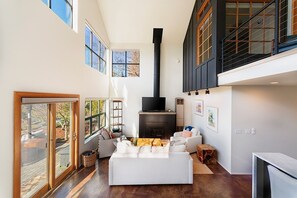 A spacious living room with a fireplace, large windows, and a high ceiling.