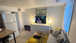 A modern living room with a sofa, a coffee table, a yellow ottoman, a TV, and a dining area with a table and chairs. - Bluffs suite retreat - beautiful cliffcrest leafy suburb moments from the beach! (Toronto)