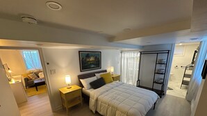A bedroom with a bed, bedside tables, a nightstand, a painting on the wall, a window with curtains, and a bathroom with a shower and a towel rack. - Bluffs suite retreat - beautiful cliffcrest leafy suburb moments from the beach! (Toronto)