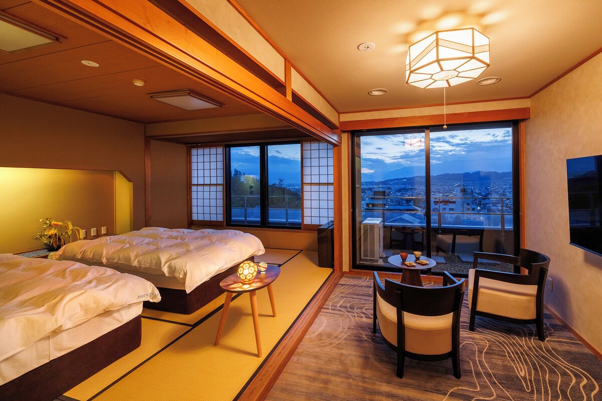 A hotel room with a bed, a small table with a lamp, a chair, and a view of the city through large windows.