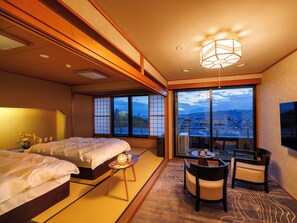 A hotel room with a bed, a small table with a lamp, a chair, and a view of the city through large windows. - SANEIKAKU (Matsumoto)
