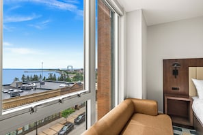 A hotel room with a large window, a bed, a nightstand, and a view of the cityscape and water.