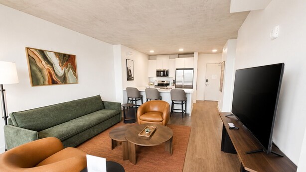 A modern living room with a sofa, chairs, a coffee table, a TV, and a kitchen area in the background. - One Bedroom King bedded room with direct views of Lake Superior (Duluth)