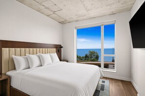 A bedroom with a large bed, a headboard, a nightstand, a window with a view of the sea, and a ceiling with a textured finish. - Large Corner 2 Bedroom/2 Bath Unit with Floor to Ceiling Lake Superior Views (Duluth)