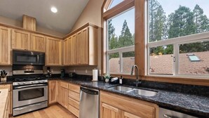 A kitchen with wooden cabinets, stainless steel appliances, a large window with a view of trees, and a skylight. - Chalet di Montagna ~ Spacious house in the pines (Mi-Wuk Village)