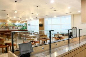 Eine großzügige Cafeteria mit Holztischen und -stühlen, einer langen Theke mit einem Computer und großen Fenstern für natürliches Licht.