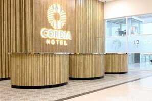A hotel reception area with a wooden reception desk and a sign that reads 'CORDIA HOTEL'.