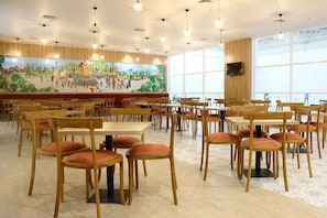 A spacious dining area with wooden tables and chairs, a mural on the wall, and large windows. - Cordia Banjarmasin (Banjarbaru)