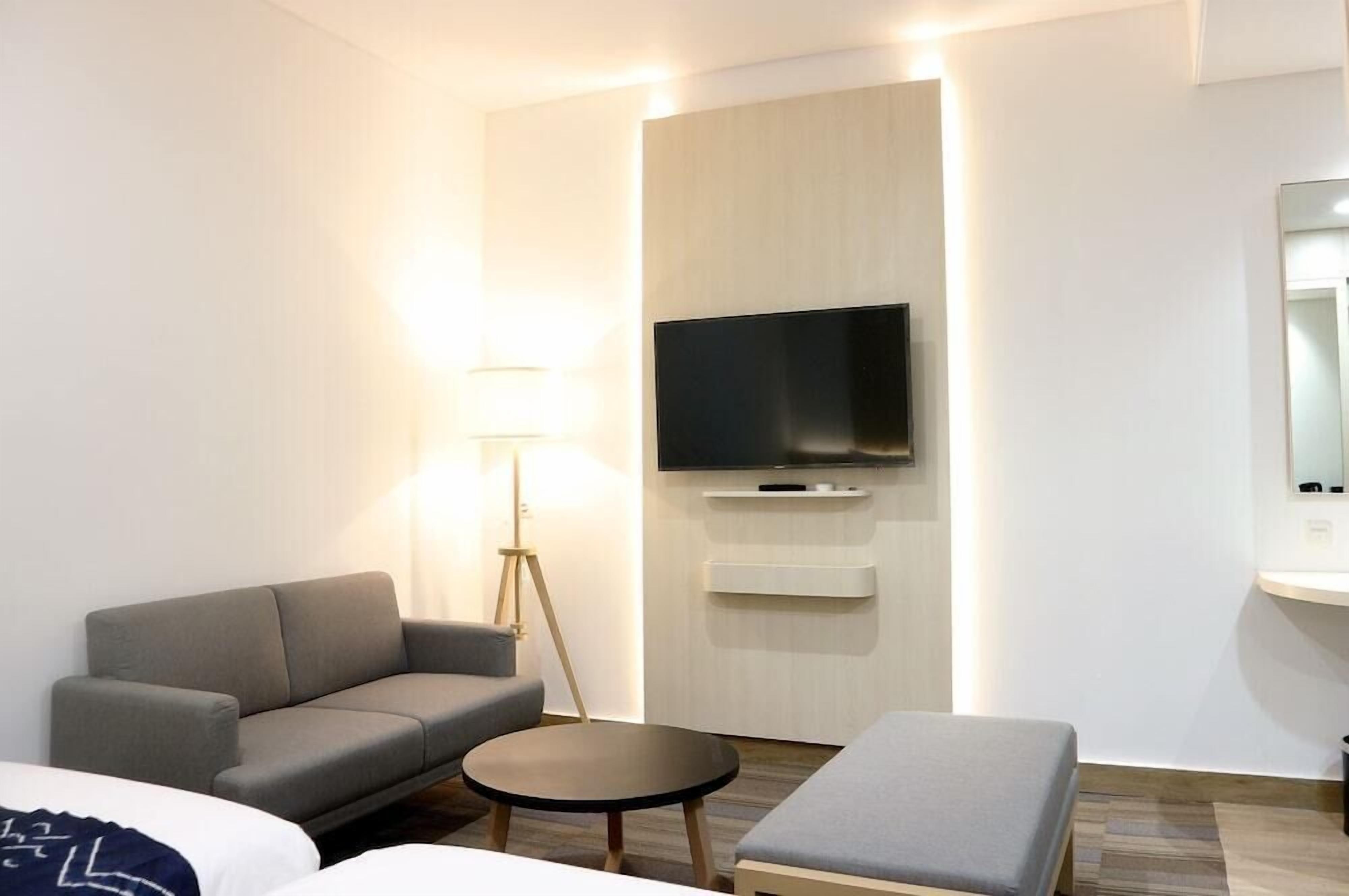 A modern hotel room with a sofa, a small round table, a flat-screen TV mounted on the wall, and a bedside table.
