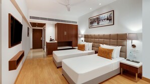 A hotel room with two beds, a TV, a ceiling fan, a painting, and a kitchenette.