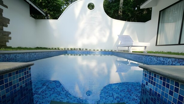 A swimming pool with blue mosaic tiles, surrounded by a white wall with a circular window and a small outdoor seating area. - Mayas Beach House Boutique Serviced Villa (Ambalapuzha)